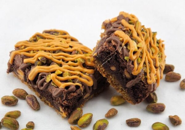 Two chocolate brownies with peanut butter drizzle and scattered pistachios around them.