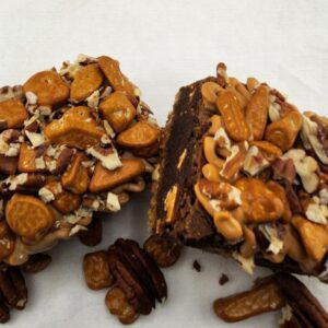 Chocolate brownies covered with nuts and pretzels, sliced in half, on a white surface.