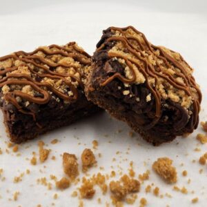 Two chocolate brownies with caramel drizzle and crumbled toppings on a white surface.