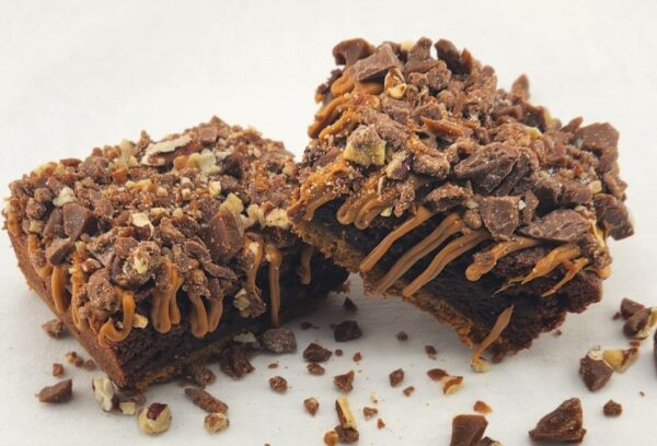 Two caramel drizzled brownies topped with chopped nuts and chocolate pieces.